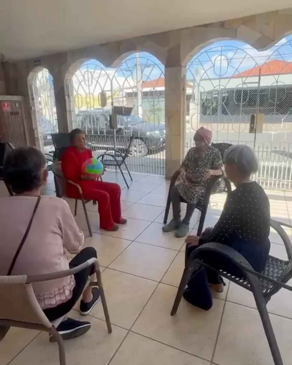 Residentes sentadas em roda na varanda coberta, passando uma bola colorida durante a atividade