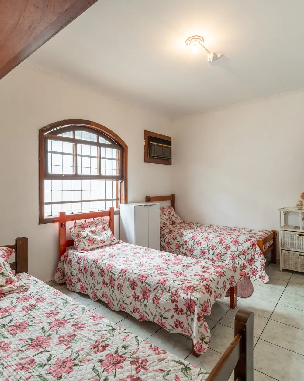 Quarto com três camas de colchas floridas, janela em arco e armários de madeira
